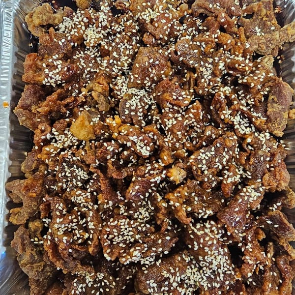 Crispy sesame beef in a foil tray, glistening with sauce and sprinkled with white sesame seeds.