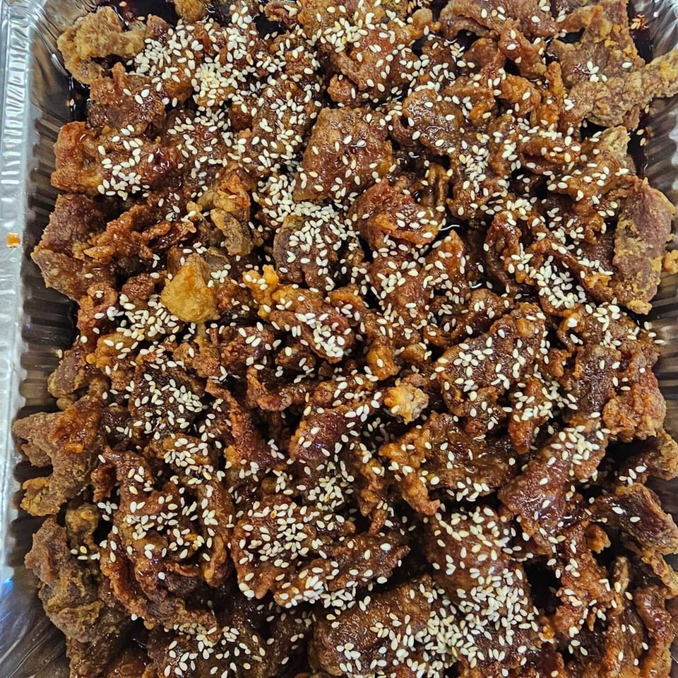 Crispy sesame beef in a foil tray, glistening with sauce and sprinkled with white sesame seeds.